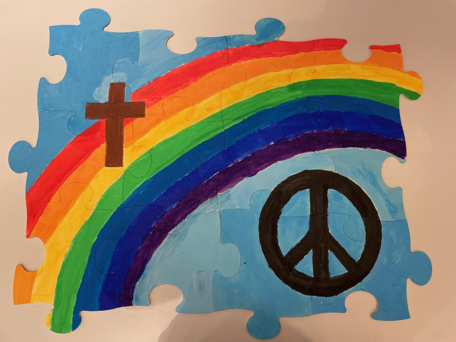Verschiedene Puzzleteile erkennbar. Der Hintergrund ist hellblau. In der Mitte des Puzzles ein Regenbogen. Oben links ein braunes Kreuz. Unten rechts das Peace-Zeichen. Das Bild wurde von den Firmlingen der Pfarrei St. Martin gemalt.