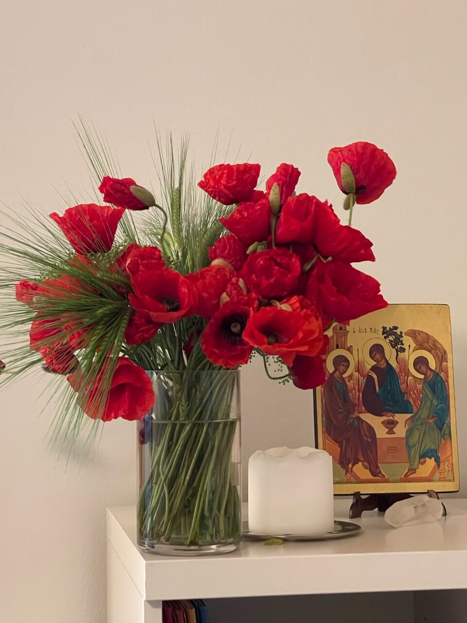 Ein weisses Sideboard. Darauf steht eine Engelsikone, eine weisse Kerze und eine Blumenvase voller Mohnblumen.