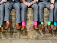 5 Männer auf einer Steinmauer. Man sieht nur die Beine der Männer. Sie sind elegant gekleidet. Alle in grau. Doch die Socken der Männer sind bunt. Alles Neonfarben.sind -- B